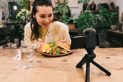 Imagen de una mujer sentada con alimentos en una mesa y mirando a la cámara colocada sobre un pequeño tripié