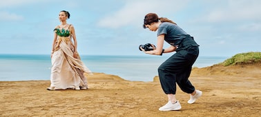 Imagen de uso en la que se muestra a una mujer con una cámara a mano que hace capturas a otra mujer con un traje de época y la costa de fondo