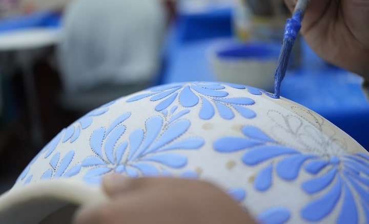 Imagen que muestra a un artesano agregando patrones a una jarra con un pincel y pintura azul. El enfoque está en el pincel, y el primer plano de la jarra y el fondo están desenfocados